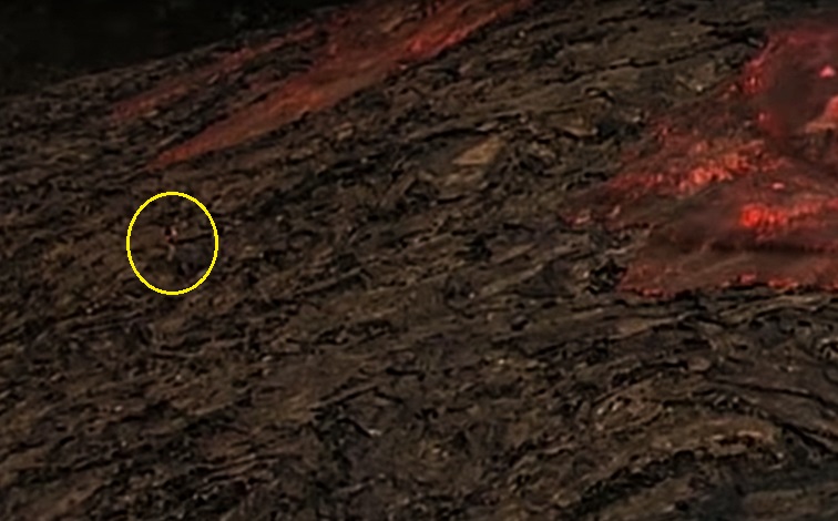 Image shows a slope of fresh lava, with two streams of red lava flowing down. A tiny, indistinct person stands between them.