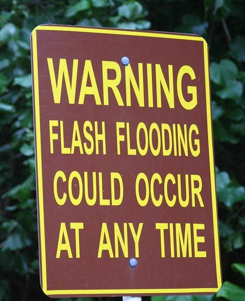 Image shows a brown warning sign with yellow lettering reading "Warning: Flash Flooding Could Occur at Any Time"